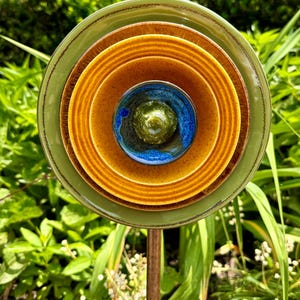 May include: A decorative garden art piece featuring stacked ceramic plates in shades of green, brown, and yellow. The center plate is blue and green. The plates are mounted on a copper rod, creating a unique outdoor sculpture.