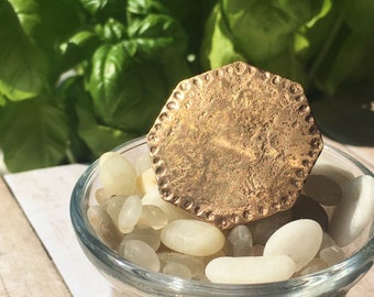 May include: A gold-toned, octagonal-shaped pendant with a textured surface sits on top of a small glass bowl filled with white stones.