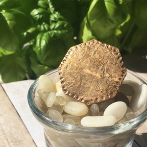 May include: A gold-toned, octagonal-shaped pendant with a textured surface sits on top of a small glass bowl filled with white stones.