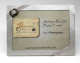 May include: A silver metal photo frame with a gray ribbon and an antique buckle. The frame is designed to hold a 5 x 7 inch photograph. The text "Antique Buckle Photo Frame" and "5 x 7 Photograph" is printed on the frame. The text "Provenance Creations Metal Detecting Finds... Transformed" and "www.ProvenanceStore.Etsy.com" is also printed on the frame.