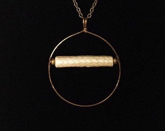 May include: A gold chain necklace with a white patterned cylinder pendant hanging from a circular wire frame.