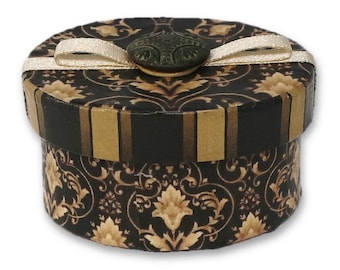 May include: A small round gift box with a black and gold patterned design. The box has a white ribbon and a decorative button.