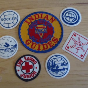 May include: A collection of vintage fabric patches with various designs and text. Includes a soccer patch, a softball patch, and patches with text such as "Indian Guides" and "Life Saving Junior". The patches are in various shapes and colors.