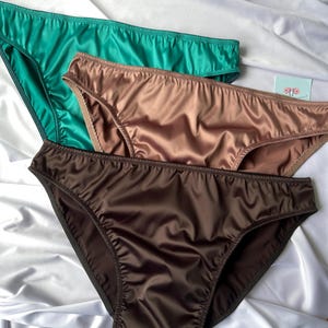 Puede incluir: Tres bragas de sat&eacute;n en tonos verde azulado, bronce y marr&oacute;n chocolate est&aacute;n dispuestas sobre una superficie de sat&eacute;n blanco. Las bragas tienen un corte cl&aacute;sico con delicados detalles de costura, mostrando un acabado suave y brillante.