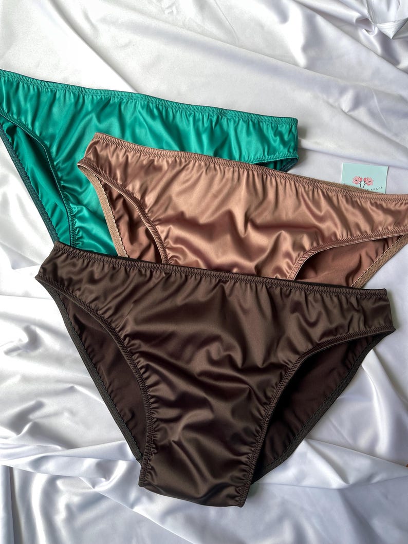 Puede incluir: Tres bragas de sat&eacute;n en tonos verde azulado, bronce y marr&oacute;n chocolate est&aacute;n dispuestas sobre una superficie de sat&eacute;n blanco. Las bragas tienen un corte cl&aacute;sico con delicados detalles de costura, mostrando un acabado suave y brillante.