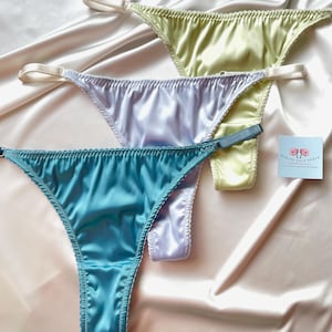 Może przedstawiać: Trzy satynowe stringi w pastelowych kolorach: jasnoniebieskim, lawendowym i żółto-zielonym. Każdy string ma cienkie paski i delikatne szwy. Bielizna ułożona jest na jedwabistej, kremowej tkaninie.
