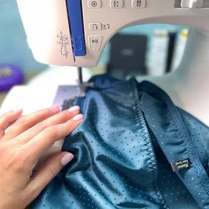 Puede incluir: Primer plano de una m&aacute;quina de coser blanca y azul cosiendo tela turquesa con un estampado de lunares. La mano de una persona sujeta la tela. La tela tiene una etiqueta que dice "Marine Eau Space".