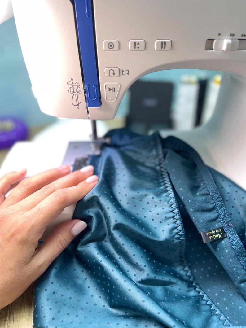 Puede incluir: Primer plano de una m&aacute;quina de coser blanca y azul cosiendo tela turquesa con un estampado de lunares. La mano de una persona sujeta la tela. La tela tiene una etiqueta que dice "Marine Eau Space".