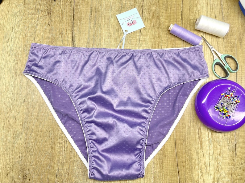 Puede incluir: Un par de ropa interior de color lavanda con ribete blanco. La ropa interior tiene un estampado de lunares y est&aacute; hecha de un material brillante. Suministros de costura, incluyendo tijeras, alfileres e hilo, est&aacute;n junto a la ropa interior.
