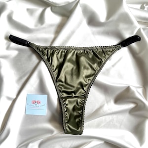 May include: Olive green satin thong underwear with black trim and adjustable straps. The lingerie is displayed on a white satin fabric. A small blue card with text and a floral design is in the lower left corner.