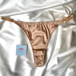 May include: A pair of beige satin thong underwear with adjustable straps. The lingerie is displayed on a white satin fabric. A small blue tag with text and a floral design is in the background.