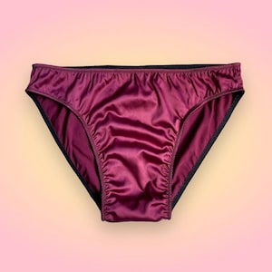 Puede incluir: Bragas de sat&eacute;n burdeos con ribete negro. La ropa interior tiene un corte cl&aacute;sico y una textura suave y brillante. El fondo es un degradado de rosa y amarillo.