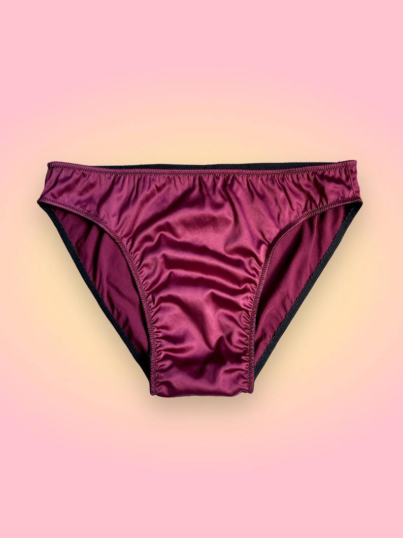 Puede incluir: Bragas de sat&eacute;n burdeos con ribete negro. La ropa interior tiene un corte cl&aacute;sico y una textura suave y brillante. El fondo es un degradado de rosa y amarillo.