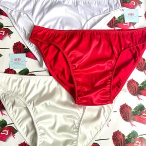 Puede incluir: Tres pares de ropa interior de sat&eacute;n en blanco y rojo. La ropa interior se muestra sobre una superficie estampada con rosas y cajas de regalo. La tela de sat&eacute;n tiene un ligero brillo y la ropa interior tiene un corte cl&aacute;sico.