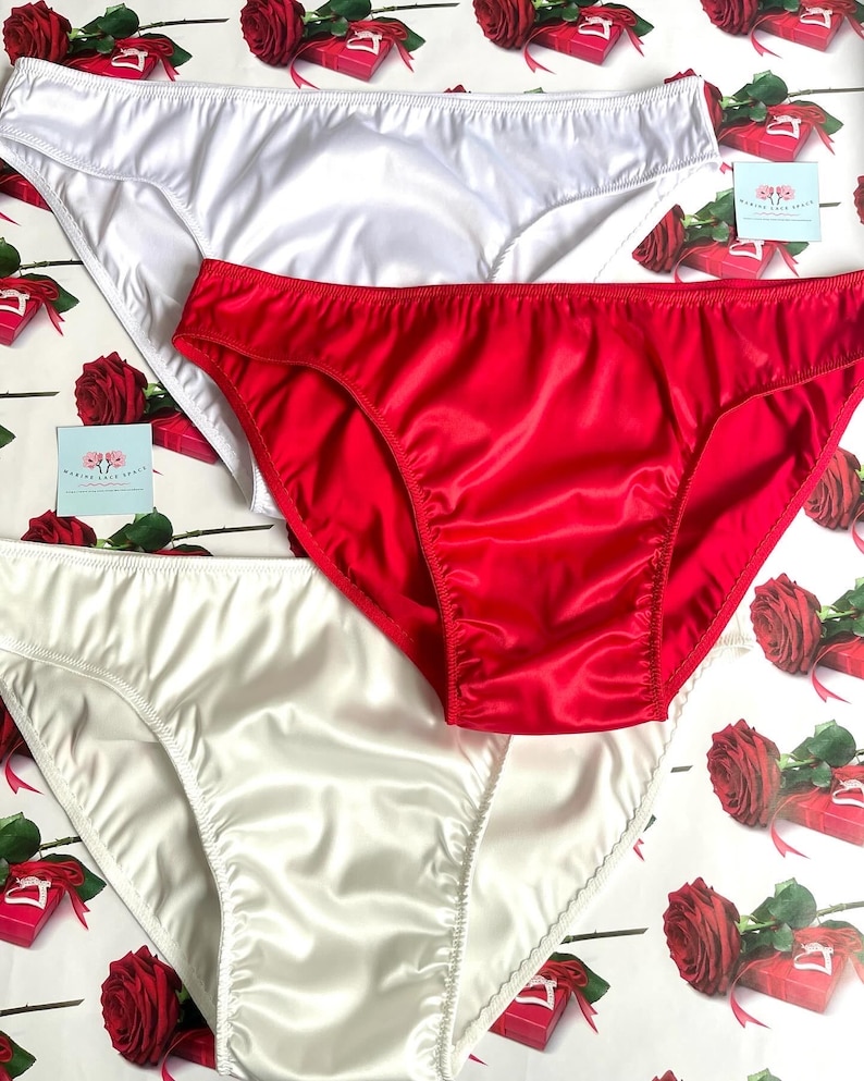 Puede incluir: Tres pares de ropa interior de sat&eacute;n en blanco y rojo. La ropa interior se muestra sobre una superficie estampada con rosas y cajas de regalo. La tela de sat&eacute;n tiene un ligero brillo y la ropa interior tiene un corte cl&aacute;sico.
