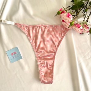 May include: A pink satin thong with white polka dots and white trim. The thong has a delicate design. A sprig of pink roses and a small card with text are in the background.