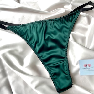 Puede incluir: Tanga de satén verde esmeralda con ribete y tirantes negros. La lencería se muestra sobre un fondo de satén blanco. Una pequeña tarjeta con un logotipo floral está en el fondo.