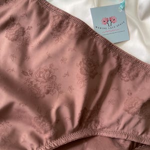 Puede incluir: Primer plano de ropa interior de color malva con un estampado floral. La tela tiene un brillo sutil y el dise&ntilde;o incluye motivos de rosas detallados. Una etiqueta con el texto "MARINE LACE SPACE" est&aacute; sujeta con una cinta azul claro.