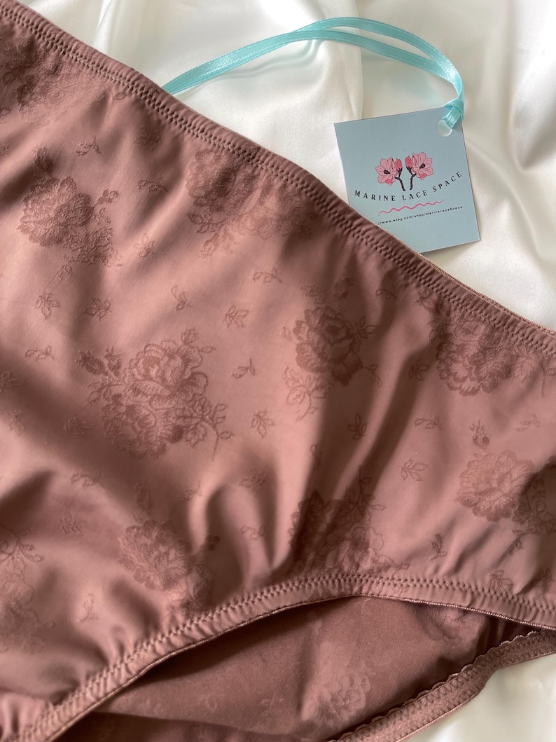 Puede incluir: Primer plano de ropa interior de color malva con un estampado floral. La tela tiene un brillo sutil y el dise&ntilde;o incluye motivos de rosas detallados. Una etiqueta con el texto "MARINE LACE SPACE" est&aacute; sujeta con una cinta azul claro.
