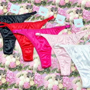 Könnte beinhalten: Fünf Paar Satin-Dessous in Schwarz, Rot, Rosa, Hellrosa und Weiß. Die Dessous sind auf einem Hintergrund aus rosa und weißen Blumen angeordnet.