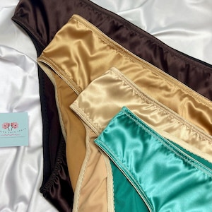 Puede incluir: Primer plano de ropa interior de sat&eacute;n en varios colores: marr&oacute;n, dorado, beige y turquesa. Las prendas est&aacute;n dispuestas sobre un fondo de sat&eacute;n blanco, resaltando su textura suave y brillo. Se ve una peque&ntilde;a tarjeta con texto.
