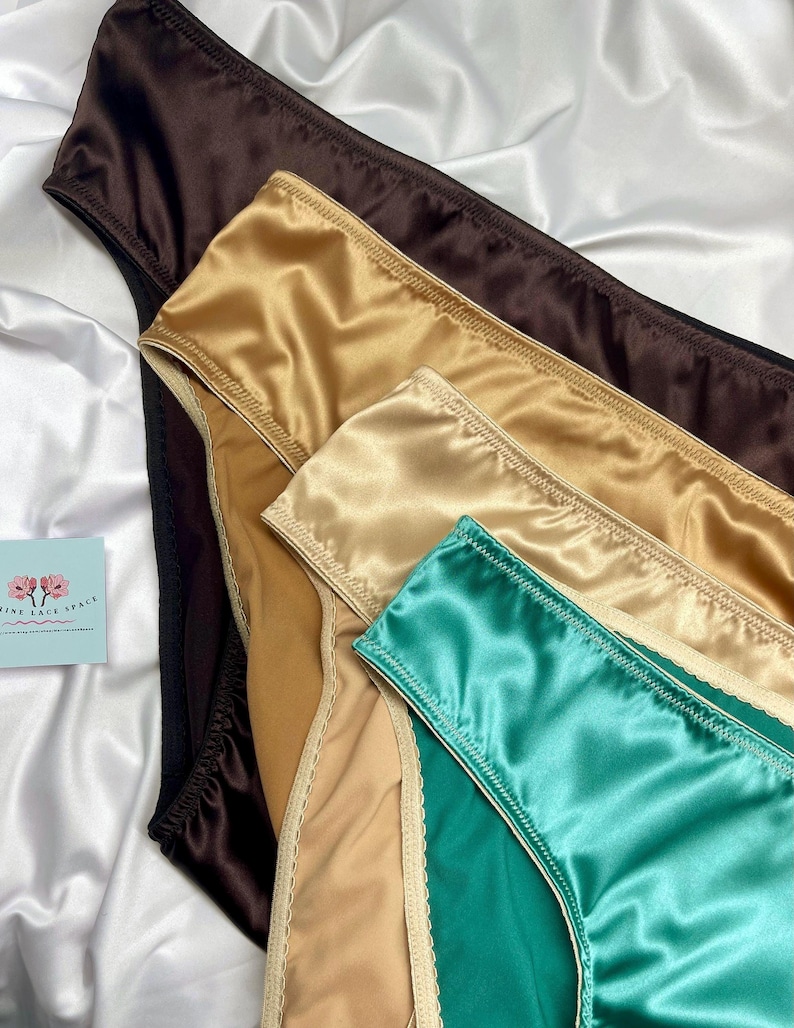 Puede incluir: Primer plano de ropa interior de sat&eacute;n en varios colores: marr&oacute;n, dorado, beige y turquesa. Las prendas est&aacute;n dispuestas sobre un fondo de sat&eacute;n blanco, resaltando su textura suave y brillo. Se ve una peque&ntilde;a tarjeta con texto.