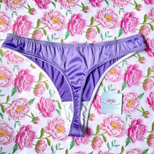 Peut inclure: Lingerie en satin violet avec bordure en dentelle blanche. La lingerie est photographiée sur un fond blanc à motif floral.