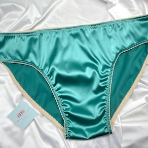 Puede incluir: Bragas de sat&eacute;n color turquesa con ribete color crema. La lencer&iacute;a est&aacute; hecha de una tela brillante y suave y se exhibe sobre un fondo de sat&eacute;n blanco. Se puede ver una peque&ntilde;a tarjeta con un dise&ntilde;o floral.