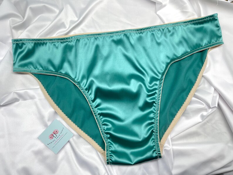 Puede incluir: Bragas de sat&eacute;n color turquesa con ribete color crema. La lencer&iacute;a est&aacute; hecha de una tela brillante y suave y se exhibe sobre un fondo de sat&eacute;n blanco. Se puede ver una peque&ntilde;a tarjeta con un dise&ntilde;o floral.