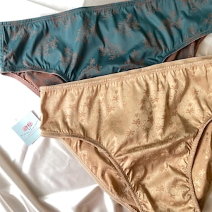 Puede incluir: Dos pares de bragas. Uno es turquesa con un estampado floral marr&oacute;n, el otro es beige con un estampado de remolino. La ropa interior est&aacute; hecha de un material sedoso y se exhibe sobre una superficie de sat&eacute;n blanco.