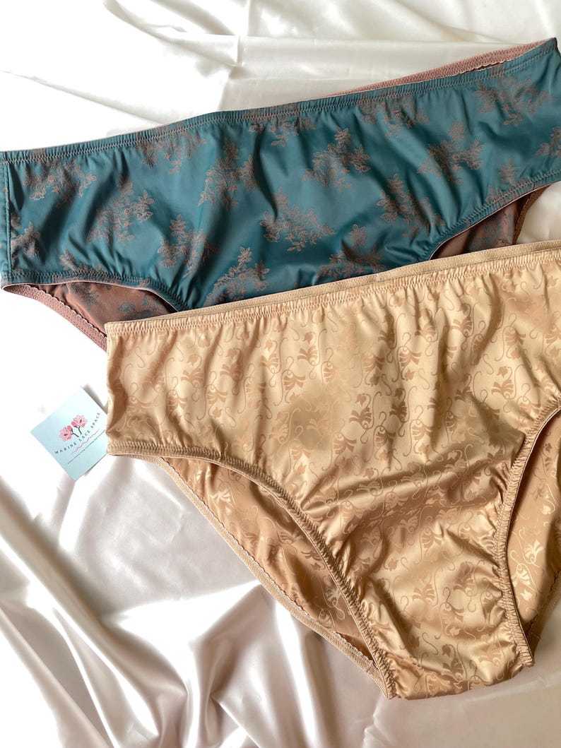 Puede incluir: Dos pares de bragas. Uno es turquesa con un estampado floral marr&oacute;n, el otro es beige con un estampado de remolino. La ropa interior est&aacute; hecha de un material sedoso y se exhibe sobre una superficie de sat&eacute;n blanco.