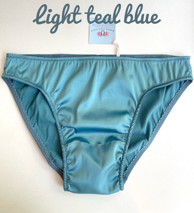 Puede incluir: Bragas de sat&eacute;n azul turquesa claro con un ribete de encaje festoneado. La etiqueta dice "MARINE LACE SPACE". El texto "light teal blue" est&aacute; arriba.