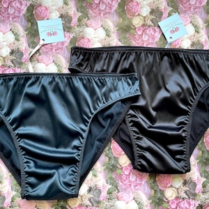 Puede incluir: Dos pares de ropa interior de sat&eacute;n negro. Un par es de color verde azulado oscuro y el otro es negro. Ambos pares tienen un dise&ntilde;o de cintura alta y se muestran sobre un fondo floral. Cada par tiene una etiqueta adjunta.