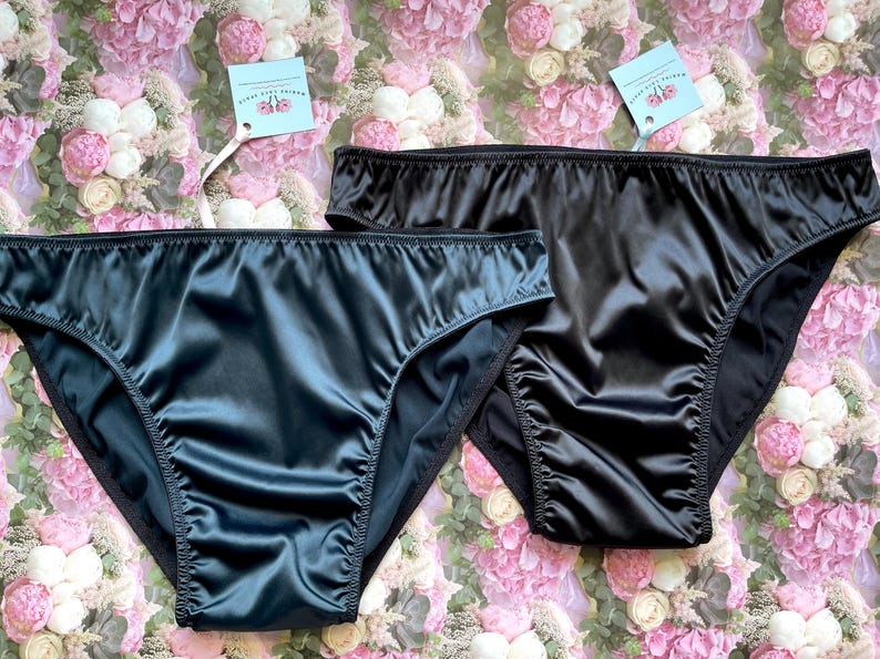 Puede incluir: Dos pares de ropa interior de sat&eacute;n negro. Un par es de color verde azulado oscuro y el otro es negro. Ambos pares tienen un dise&ntilde;o de cintura alta y se muestran sobre un fondo floral. Cada par tiene una etiqueta adjunta.