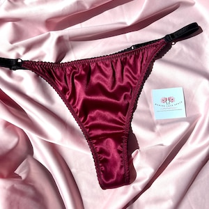 Może przedstawiać: Bordowy satynowy string z czarnym wykończeniem i ramiączkami. Bielizna jest prezentowana na różowej satynowej tkaninie. Mała biała karta z napisem "MARINE LACE SPACE" znajduje się w prawym dolnym rogu.