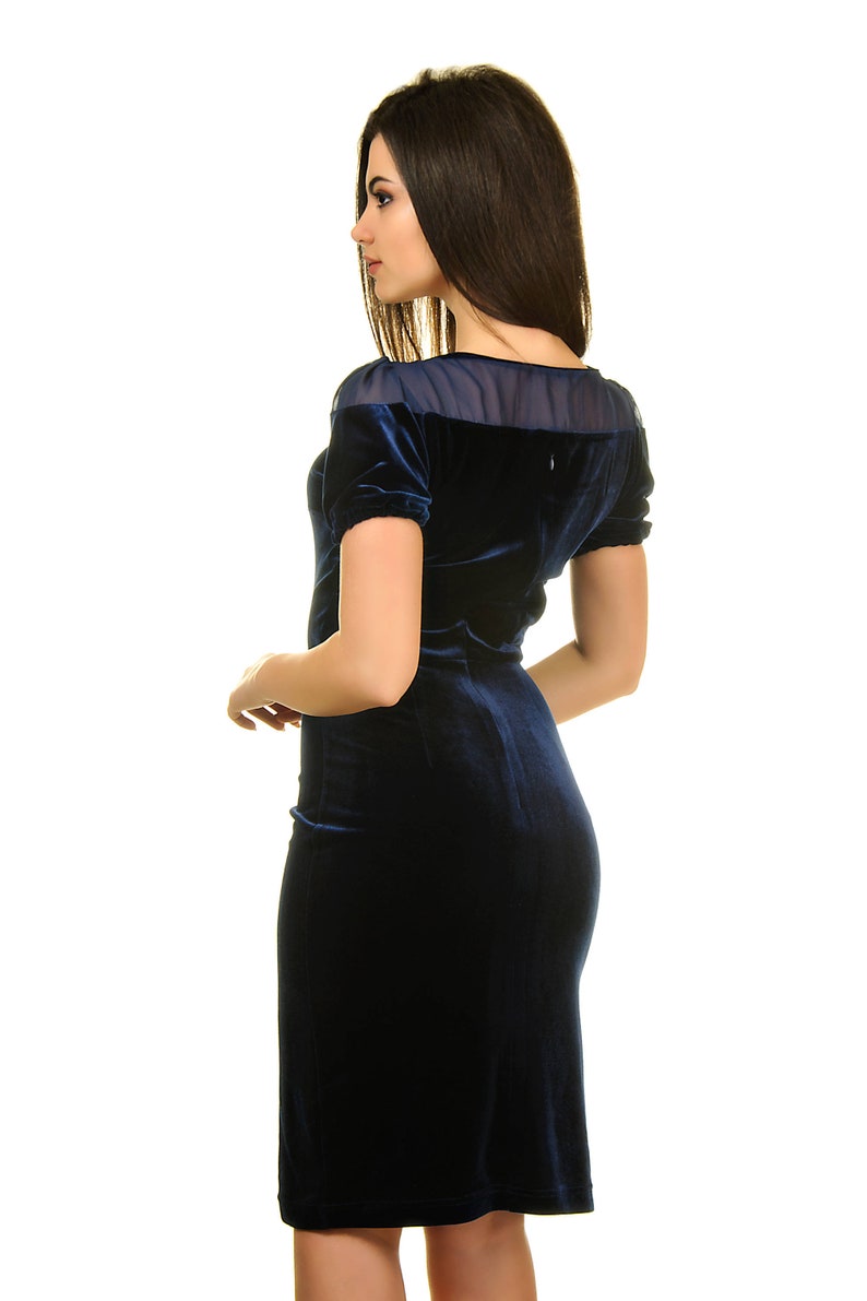 May include: A woman wearing a dark blue velvet dress with a sheer overlay at the neckline. The dress has short sleeves and a fitted silhouette.