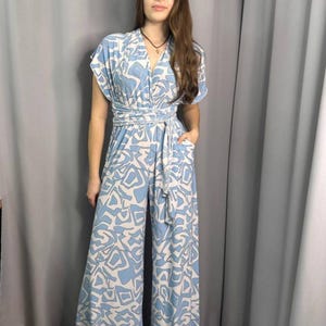 May include: A light blue jumpsuit with a white abstract pattern. The jumpsuit has a V-neck, short sleeves, and a wide leg. It features a tie belt at the waist.