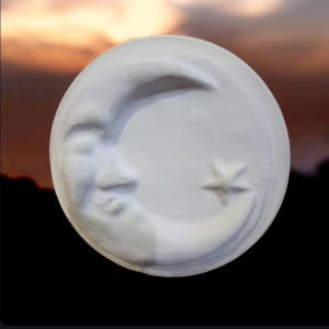 May include: A white, circular ceramic piece featuring a crescent moon with a smiling face and a five-pointed star. The moon and star are slightly raised from the surface. The background is a blurred gradient of orange, yellow, and black.