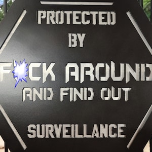 May include: A black hexagonal sign with silver lettering that reads "Protected by F*ck Around and Find Out Surveillance".