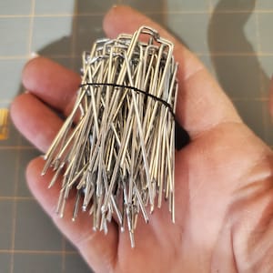 May include: A close-up of a bundle of silver metal staples. The staples are arranged in a vertical stack and are held together by a black rubber band.