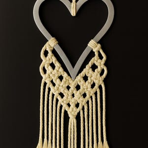 May include: A heart-shaped wall hanging featuring a silver metal frame and cream-colored macrame detailing. The macrame includes a woven pattern and fringe. The piece is suspended by a cream-colored cord, creating a decorative accent.