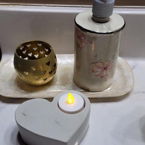 May include: A white heart-shaped candle holder with a small, round, white candle with a yellow flame inside. The candle holder is on a white countertop next to a floral patterned soap dispenser and a brass candle holder.