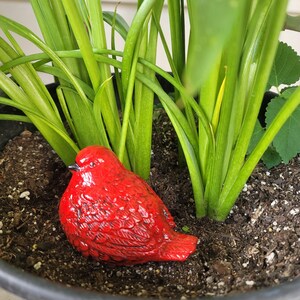 May include: A vibrant red ceramic bird figurine rests in a black plant pot filled with soil and surrounded by green plant leaves. The bird has detailed feather textures and a small black beak. The plant pot is made of plastic.