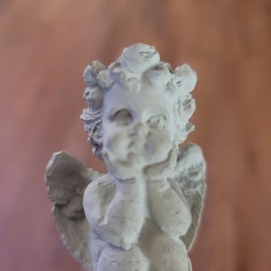 May include: A white ceramic cherub figurine with wings, holding its hands to its face. The cherub has a sweet expression and is a decorative piece.