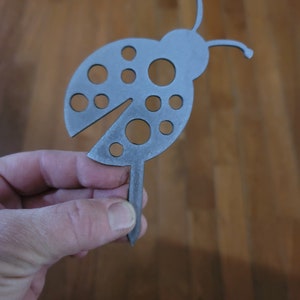 May include: A metal ladybug garden stake with a cut-out design and a pointed end for inserting into the ground. The ladybug is silver in color and has black dots.