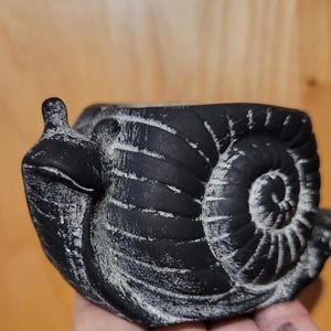 May include: Black and white snail-shaped planter with a textured finish. The planter is made of ceramic and has a wide opening for planting.
