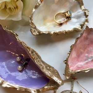 K&ouml;nnte beinhalten: Drei dekorative Austernmuschel-Schalen mit goldenen Akzenten. Die obere Schale ist wei&szlig; mit einem goldenen Ring im Inneren. Die mittlere Schale ist lavendelfarben mit goldenen Akzenten und einem Paar Perlenohrringen im Inneren. Die untere Schale ist rosa mit goldenen Akzenten und einem Paar Kristallohrringen im Inneren.