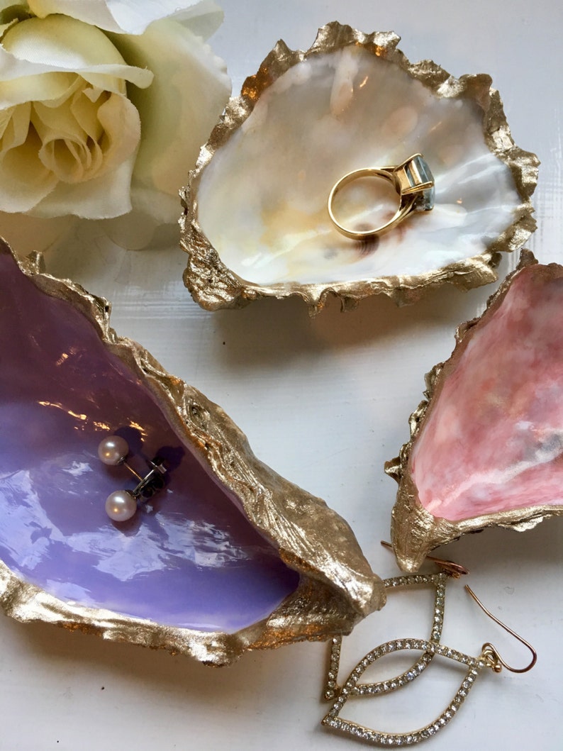 K&ouml;nnte beinhalten: Drei dekorative Austernmuschel-Schalen mit goldenen Akzenten. Die obere Schale ist wei&szlig; mit einem goldenen Ring im Inneren. Die mittlere Schale ist lavendelfarben mit goldenen Akzenten und einem Paar Perlenohrringen im Inneren. Die untere Schale ist rosa mit goldenen Akzenten und einem Paar Kristallohrringen im Inneren.