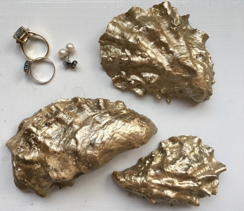 K&ouml;nnte beinhalten: Drei goldbemalte Austernmuscheln auf wei&szlig;em Hintergrund. Die Muscheln sind in verschiedenen Gr&ouml;&szlig;en und Formen, mit einer rauen, strukturierten Oberfl&auml;che.