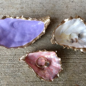 K&ouml;nnte beinhalten: Drei Ringteller aus Austernmuscheln, jede in einer anderen Farbe bemalt. Die erste ist lila, die zweite wei&szlig; und die dritte rosa. Jede Schale hat einen goldenen Rand und ist mit einem goldenen Maldesign verziert.